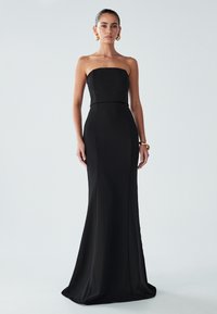 Nainen, jolla on päällä hihaton musta lantionpituinen mekko, jossa on tiukka yläosa, sekä kultaketjut ja rannekorut, seisoo yksinkertaista taustaa vasten.