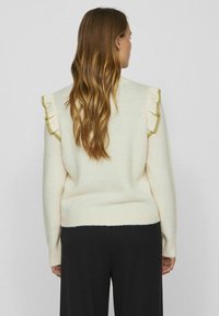 Kremowy sweter z dzianiny z falbankowymi akcentami na ramionach w kontrastowym wykończeniu, uniwersalny krój i teksturowana powierzchnia, noszony z ciemnymi spodniami.