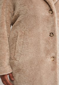 Manteau long, beige et en velours, confectionné dans un matériau doux et pelucheux. Il présente un col en V, quatre boutons et une poche avant.