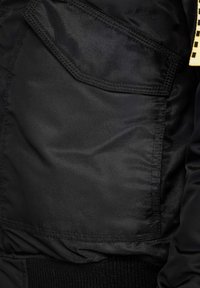 Svart bomberjacka med en slät, något glänsande textur. Har en stor sidficka och en ribbad midja. Minimalistisk design.