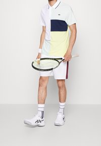 Kortärmad polo i ljusblått, marinblått och gult, tillsammans med vita shorts. Håller en tenniskejd. Vita sportskor och randiga strumpor.