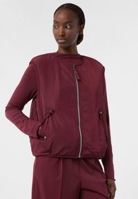 Burgundowa pikowana kamizelka z okrągłym dekoltem, z przodu na zamek błyskawiczny i bocznymi kieszeniami, noszona na długim rękawie. Gładka faktura, minimalistyczny design.