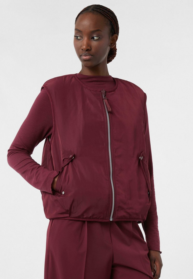 Burgundowa pikowana kamizelka z okrągłym dekoltem, z przodu na zamek błyskawiczny i bocznymi kieszeniami, noszona na długim rękawie. Gładka faktura, minimalistyczny design.