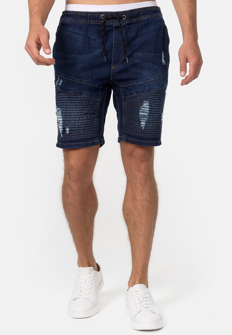 Shorts en denim bleu foncé avec une taille à cordon de serrage, présentant des accents côtelés et des détails usés. Ils ont une coupe ajustée et des bords ourlés.