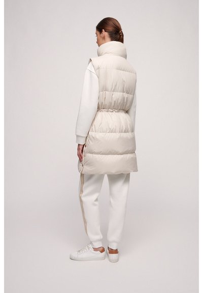 Beige Steppweste mit hohem Kragen, tailliertem Schnitt, gestepptem Muster und seitlichen Knöpfen, getragen über einem weißen Sweatshirt und Jogginghosen.
