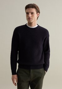 Marine gebreide trui met een gestructureerd patroon en geribde manchetten en zoom, gecombineerd met olijfgroene broek. Ronde halslijn en lange mouwen.