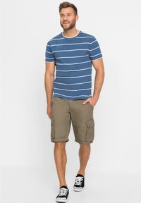 T-shirt a righe blu con strisce bianche, abbinata a pantaloni cargo beige con tasche laterali. Indossata con sneakers nere e bianche.