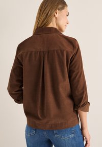 Camicia di velluto a coste marrone con colletto, polsini abbottonati e dettagli plissettati sulla schiena. Abbinata a jeans blu, presenta una texture morbida e una vestibilità su misura.