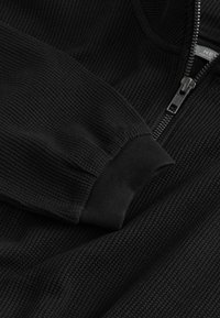 Černá rozepínací mikina s ribovanou texturou, s vysokým límcem a elastickými manžetami. Na přední straně je viditelný zip a detail loga.
