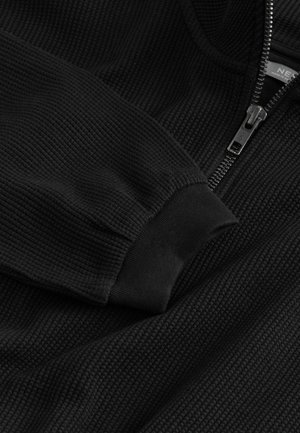 Sort zip-up sweatshirt med ribbet tekstur, der har en høj krave og elastiske manchetter. Lynlås og logo detaljer synlige på forsiden.