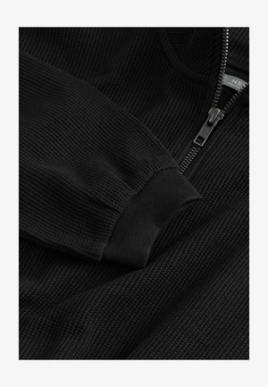 Črna zip-up sweatshirt s ribastim teksturo, ki ima visok ovratnik in elastične manšete. Zadrga in detajl logotipa sta vidna spredaj.