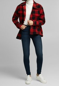 Camisa de franela a cuadros rojos y negros con botones a presión, combinada con jeans skinny de mezclilla oscura y zapatillas blancas. Textura suave, diseño casual.