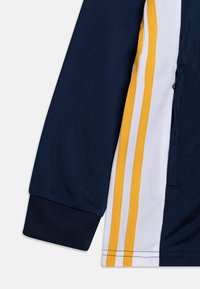 Chaqueta atlética en marino y blanco con rayas amarillas, fabricada con un tejido texturizado y un bolsillo con cremallera en el lateral. Mangas con puños.