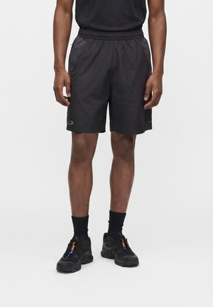 Homme portant un short de sport noir, des chaussettes noires et des baskets noires avec des tirettes de fermeture éclair colorées, debout devant un fond uni.