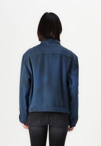 Τζάκετ τζην σε σκούρο μπλε χρώμα με χαλαρή εφαρμογή, εμφανείς ραφές και κλασικό γιακά. Φοράει με μαύρα τζιν.