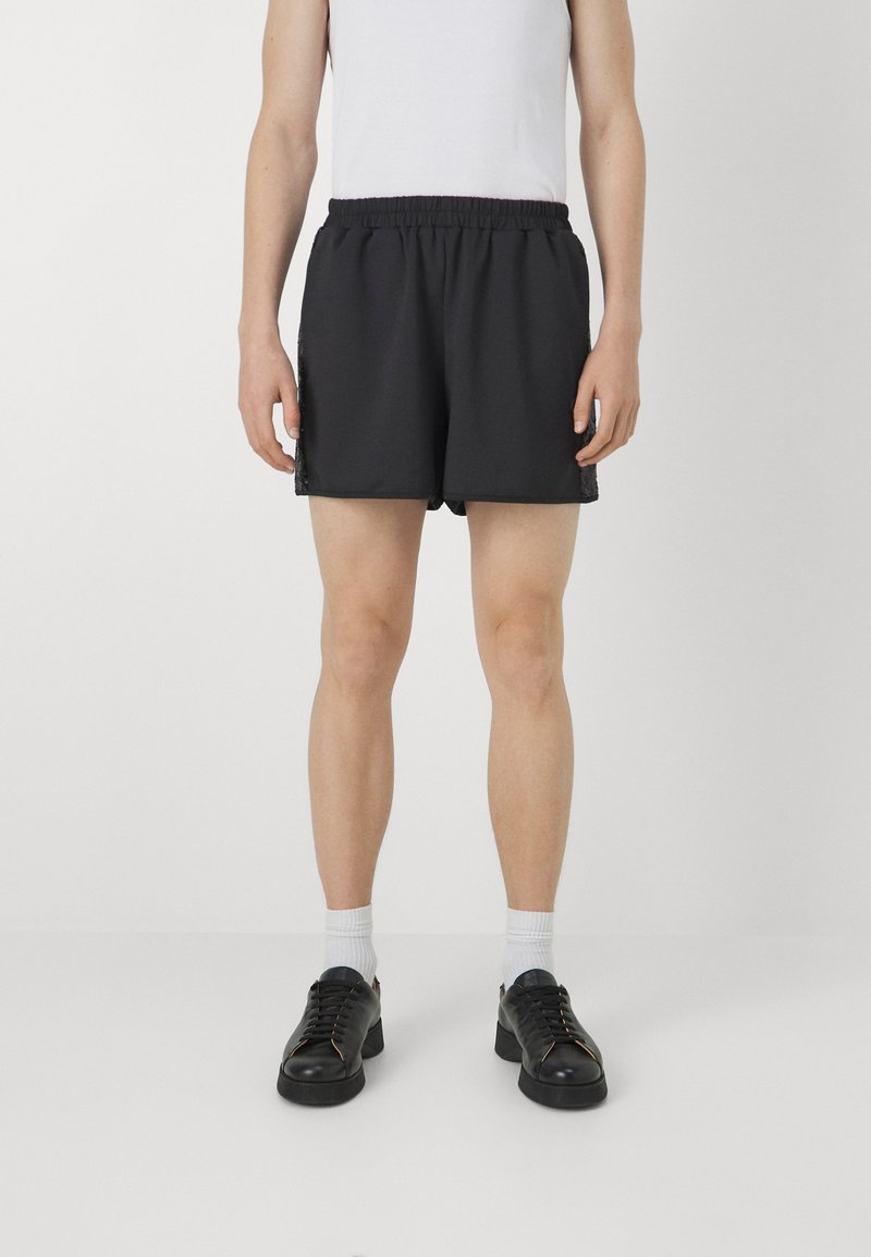 Black athletic shorts with an elastic waistband and side mesh panels, worn with a white tank top and black shoes. White socks visible.