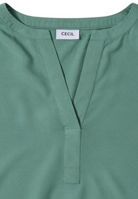 Grüne Bluse von CECIL mit V-Ausschnitt und glatter Textur. Der Stoff ist leicht und hat einen subtilen Glanz sowie saubere Nähte.