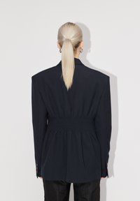 Blazer marine ajusté avec des épaules larges et une taille élastiquée. Tissu lisse et poignets à boutons. Lignes épurées et silhouette structurée.
