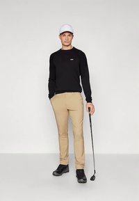 Jauns vīrietis, tērpies melnā džemperī, bēšās biksēs, baltā cepurē un melnās kurpēs, tur golfu nūju, stāvot pret vienkāršu pelēku fona.