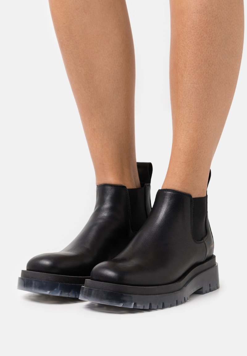 Bottes basses en cuir noir avec semelles claires à crampons portées sur des jambes nues sur un fond blanc.