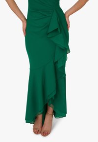 Zelené šaty s vrstvenými volány, vyrobené z hladké tkaniny. Mají přiléhavý korzet a rozšířený lem, doplněné o holé sandály na řemínky.