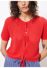 Rote gestrickte kurzärmlige Strickjacke mit einem strukturierten Rautenmuster, vorn mit Bindebändern und einem runden Halsausschnitt.