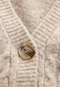 Beezvärviline kudumiste lõngaga kardigan, millel on tekstuuriline muster ja suur ümmargune marmorist nööp kaela juures.