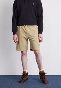 Beige shorts with side pockets and elastic waistband, paired with a black hoodie and brown boots with orange accents on a blue carpet.