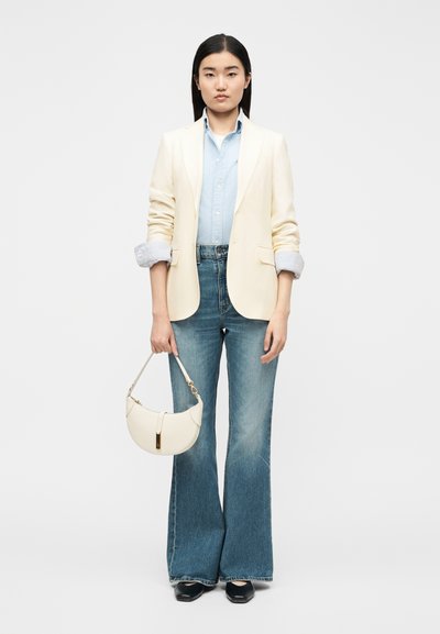 Νεαρή γυναίκα με κρεμ σακάκι, γαλάζιο πουκάμισο, τζιν παντελόνι με καμπάνες, μαύρα παπούτσια, κρατώντας μια μικρή λευκή τσάντα χειρός σε λευκό φόντο.