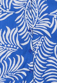 Tissu bleu avec des motifs de feuilles tropicales blanches et une rangée verticale de quatre boutons bleus assortis.