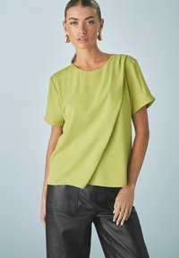 Blusa lime green a maniche corte con scollatura rotonda, texture liscia e dettagli drappeggiati, abbinata a pantaloni in pelle nera.