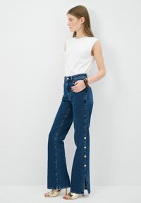 Vaqueros de mezclilla azul de tiro alto y pernera acampanada con botones laterales, combinados con una blusa sin mangas blanca y sandalias de tacón metálicas. Textura suave.