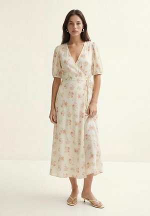 Femme debout portant une robe portefeuille crème à imprimé floral rose, manches bouffantes et sandales à talons beiges sur un fond clair uni.