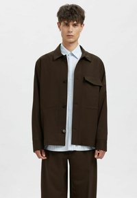 Brun jakke med lange ermer og knappelukking, med brystlomme, brukt over en lyseblå stripete skjorte og brune bukser.