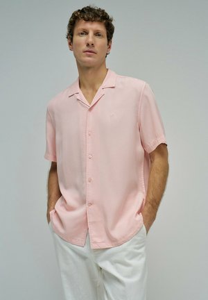 Homme portant une chemise rose clair à manches courtes boutonnée avec un pantalon blanc, debout les mains dans les poches devant un fond uni.