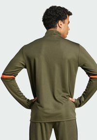 Olive green athletic long-sleeve top with a high collar, featuring a textured finish and orange accent stripes on the sleeves.
