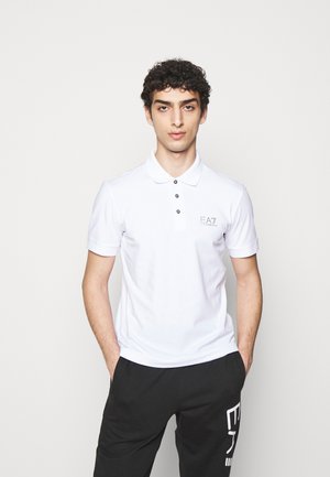 Camisa polo de algodón blanca con mangas cortas, tapeta de tres botones y logo bordado en el lado izquierdo. Combinada con pantalones negros.