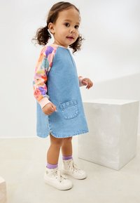 Denimjurk met kleurrijke, lange mouwen met patroon, een borstzak aan de voorkant en een afgeronde zoom. Gecombineerd met witte sneakers en paarse sokken.