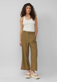 Pantaloni larghi color khaki con davanti piatto, dotati di passanti per cintura, tasche laterali e una texture del tessuto liscia. Abbinati a un top bianco senza maniche.