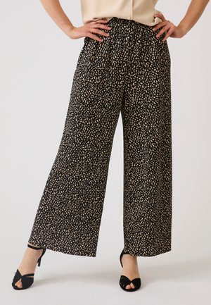 Donna che indossa pantaloni neri a gamba larga con stampa astratta beige e tacchi neri aperti sulle dita, in piedi con le mani sui fianchi su sfondo uniforme.
