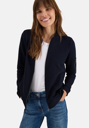 Marineblauer Bomberjacke mit gerippter Textur, Reißverschluss vorne und Seitentaschen. Über einem weißen T-Shirt getragen, kombiniert mit blauen Jeans.