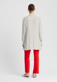Hellgraues Langarmshirt mit Rückenschlitz, kombiniert mit roten, enganliegendem Hosen und silbernen Slippern, von hinten betrachtet.