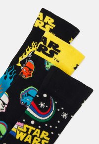 Trois paires de chaussettes noires avec des graphiques colorés de Star Wars, comprenant des stormtroopers et des planètes, avec un branding jaune en haut.