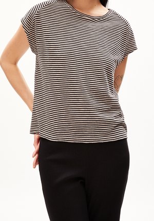 Osoba w krótkim rękawie, czarno-białej koszulce w paski i czarnych spodniach, stojąca z jedną ręką za plecami.