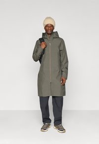 Olívazöld esőkabát elől cipzárral, kapucnival és kontrasztos cipzár részletekkel. Sötét nadrággal és bézs kötött sapkával viselve, hátizsákot cipel.