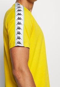 Camiseta de algodón amarilla con un patrón gráfico negro en una franja blanca a lo largo del hombro. Mangas cortas, diseño de cuello redondo, textura lisa.