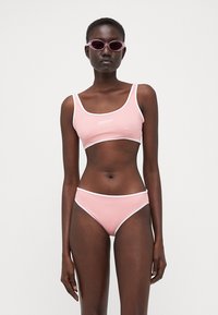 Rozā bikini komplekts ar baltu apmalīti, kurā ietilpst krope topi un zems jostas augstums apakšbikses. Gluds, elastīgs materiāls; logo uz topa priekšpuses.