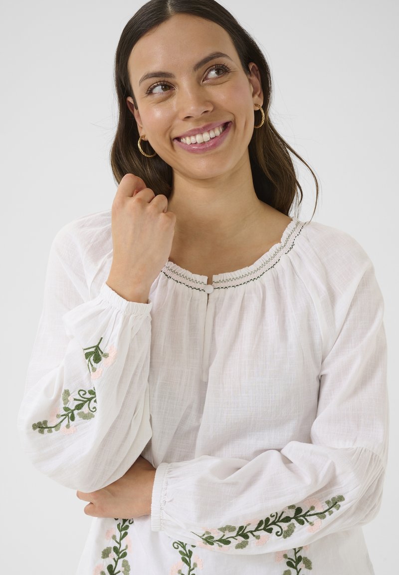 Blusa bianca di lino con maniche lunghe e puffate. Presenta ricami floreali verdi e rosa lungo le maniche e un collo arricciato con chiusura a bottone.
