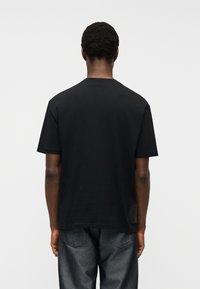 Άνδρας με κοντά σγουρά μαλλιά, φορώντας ένα απλό μαύρο κοντομάνικο μπλουζάκι και σκούρο μπλε τζιν, στέκεται γυρισμένος με την πλάτη σε απαλό φωτεινό φόντο.