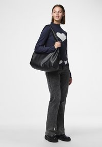 Svart skinn skulderveske med myk tekstur, stor kapasitet og enkelt design. Båret over skulderen med et avslappet antrekk bestående av en marineblå genser med hjerter.